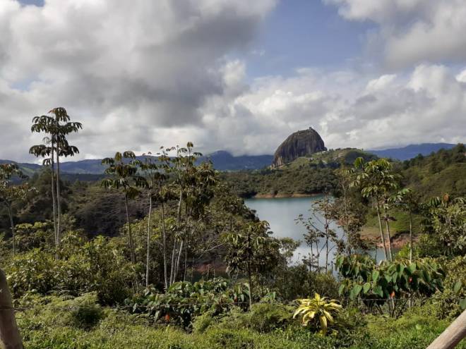 A large rock sits in the distance across a jungle landscape and a glimpse of a river in the foreground. The sky is mostly cloudy.