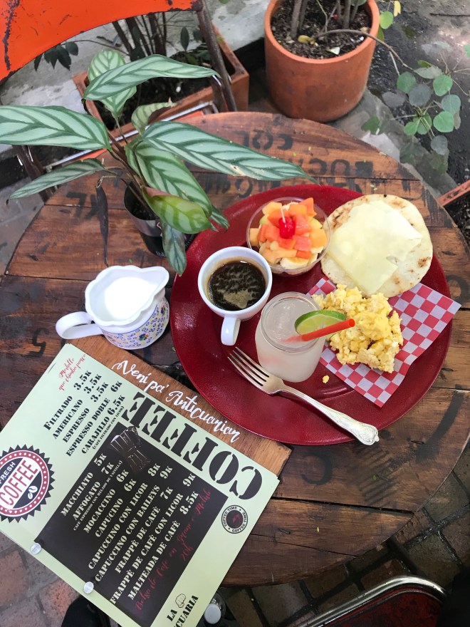 Scrambled eggs, an arepa with cheese melted on it, a cup of fruit, a coffee with a side of cream and a lemonade on a plate on a wooden cafe table with a coffee menu and a potted plant next to it.