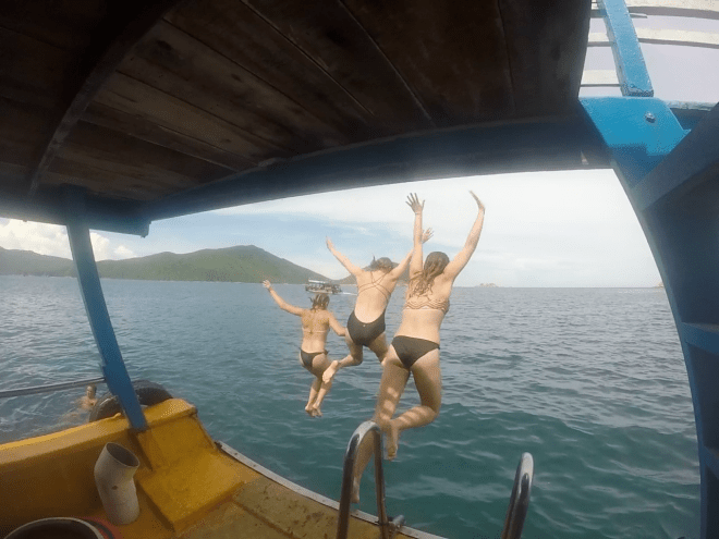 3 women in bathing suits jumping off the back of a boat into the ocean with their arms in the air.